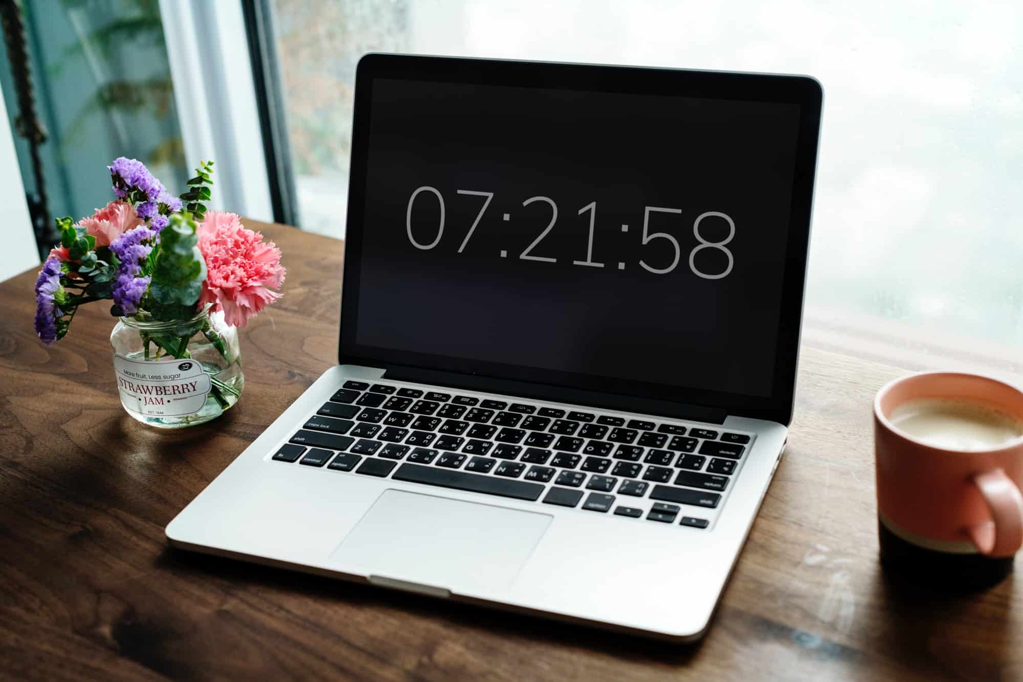 Laptop displaying time on a wooden desk with flowers and coffee.